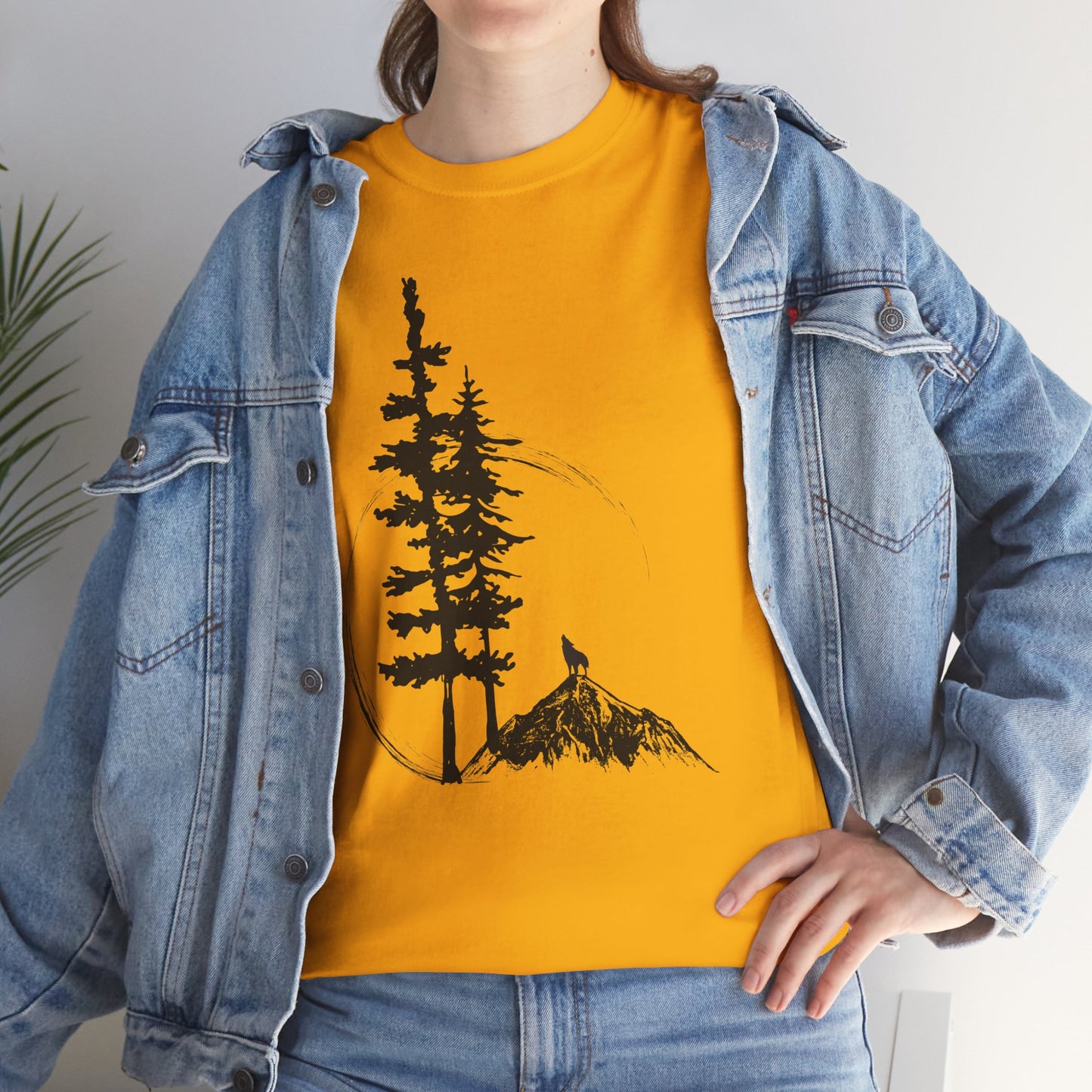 Person wearing a mustard yellow t-shirt with a tree and mountain design, paired with a denim jacket.